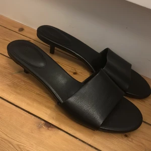 Svarta sandaler med låg klack - Stilrena svarta sandaler från H&M med öppen tå och låg klack. Skorna har en bred rem över foten och är tillverkade i syntetmaterial med en platt sula framtill. Perfekta för en trendig och minimalistisk look.