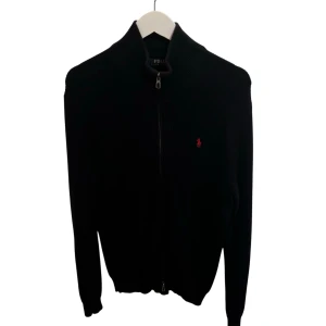 Marinblå kofta från Polo Ralph Lauren - Marinblå stickad kofta från Polo Ralph Lauren med hel dragkedja framtill och klassisk röd logga på bröstet. Hög krage och ribbade muddar ger en clean och stilren look. Perfekt för lager-på-lager under kyliga dagar.