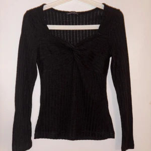 Svart ribbad långärmad topp  - Svart långärmad topp med ribbat tyg och snygg twistad detalj framtill vid bysten. Toppen har fyrkantig ringning och sitter figurnära. Perfekt att styla med jeans eller kjol för en trendig look.