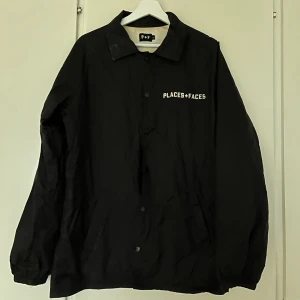 Svart vindjacka från Places+Faces - Svart vindjacka från Places+Faces med vit logga på bröstet och stort vitt kors på ryggen. Jackan har klassisk krage, knäppning framtill och två fickor. Utsidan och trycket är något sliten/fläckig och innerfodret är missfärgat.