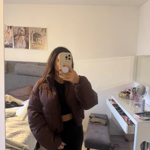 Brun pufferjacka med dragkedja - Säljer en brun pufferjacka med croppad passform och dragkedja framtill. Jackan är vadderad och har en hög krage som håller dig varm. Perfekt för kyliga dagar och ger en cool vibe till din outfit.