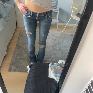 Lågmidjade blå slitna jeans - Säljer ett par blå jeans med låg midja och slitna detaljer på benen. Jeansen har klassisk femficksdesign, smal passform upptill och lätt utsvängda ben. Materialet är denim och färgen är mellanblå med slitningar för en cool look. Dom har vissa defekter, jag har själv sytt om dom lågmodjade och det är inte helt noga gjort så det är lite skrynkligt mellan benen, men inget som syns och går lätt att fixa. De har flertal hål för en sliten design. Vet dock ej vart de är från pga köpt second hand!