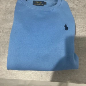 Blå tröja från Polo Ralph Lauren - Snygg blå tröja från Polo Ralph Lauren med klassisk rund hals och broderad logga på bröstet. Tröjan har lång ärm och är tillverkad i mjuk bomull, perfekt för en chill och stilren look.