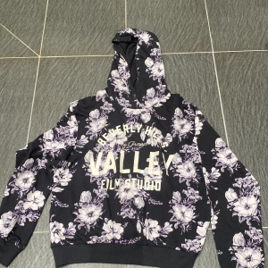 Blommig hoodie från KappAhl - Svart hoodie från KappAhl med lila och vita blommor över hela tröjan. Stor tryckt text på bröstet: 'Beverly Hills Valley Film Studio'. Mjuk bomullskänsla, huva och långa ärmar. Perfekt för dig som gillar en cool och unik stil. Har en liten fläck på sig som du ser på sista bilden men inget men lägger märke till.