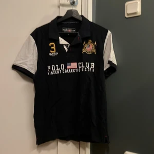 Pikétröja Polo Club Saint Vincent - Marinblå pikétröja från Polo Club Saint Vincent med vita ärmar och detaljer i gult och vitt. Broderad logga och amerikansk flagga på bröstet, samt klassisk krage och knappar. Snygg och sportig stil som sticker ut. Använd endast någon gång. Inga defekter. 