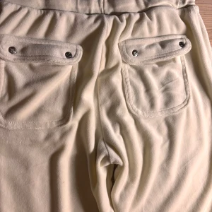 Beige mjukisbyxor med fickor och snörning - Snygga beige mjukisbyxor från Juicy Couture med bred resår i midjan, snörning och stora fickor med lock och silverfärgade knappar bak. Mjukt och skönt material, perfekt för chill dagar. Broderad JC-logga framtill på ena fickan.