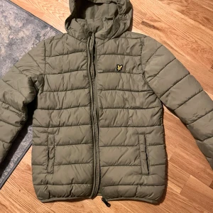 Olivgrön pufferjacka från Lyle & Scott - Snygg olivgrön pufferjacka från Lyle & Scott med huva och dragkedja framtill. Jackan har quiltad design, två fickor och den klassiska loggan på bröstet. Perfekt för kyliga dagar och enkel att matcha med det mesta.