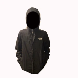 Svart vindjacka från The North Face - Svart vindjacka från The North Face med huva och dragkedja framtill. Jackan har logga på både bröstet och ryggen, samt praktiska fickor på sidorna. Tillverkad i lätt och slitstark polyester, perfekt för blåsiga dagar med ett litet håll tyvär inne från på högra fickansom inte e synligt .om man inte kollar i fickan