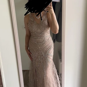 Champagnefärgad långklänning med paljetter - Säljer en magisk långklänning i champagnefärg med glittriga paljetter och pärlor i ett unikt mönster. Klänningen har v-ringning, är ärmlös och har en slits framtill samt fjäderdetaljer längst ner. Perfekt för bal eller nyår, ger riktigt lyxiga vibes!