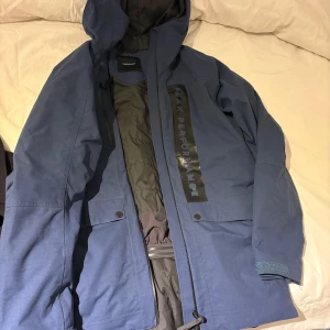 Blå parkasjacka Peak Performance - Snygg blå parkasjacka från Peak Performance med huva och stora fickor framtill. Jackan har dragkedja och tryckknappar, samt ett svart Peak Performance-tryck längs bröstet. Perfekt för kalla dagar med sitt vadderade foder och sportiga look.