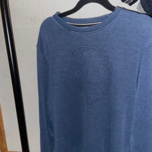 Blå Stone Island tröja i bomull - Säljer en blå långärmad tröja från Stone Island i 100% bomull. Tröjan har ett stort broderat Stone Island-logo på bröstet och en klassisk rund hals. Perfekt för dig som gillar stilrena och snygga plagg med streetkänsla.