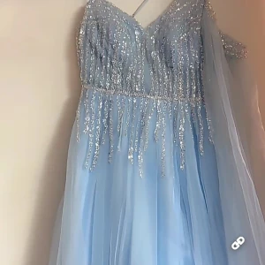 Ljusblå glittrig balklänning - Magisk ljusblå långklänning med glittriga paljettdetaljer upptill och v-ringad urringning. Klänningen har omlottkänsla, snörning i ryggen och ett svepande, tunt tyg som ger en drömmig look. Perfekt för bal eller speciella tillfällen.