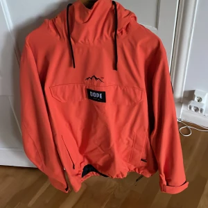 Orange skidjacka från Dope - Snygg orange skidjacka från Dope med stor huva och svarta snören. Jackan har en stor ficka framtill med Dope-logga, dragkedjor i sidorna samt i vänster ärm för att exempelvis förvara liftkort. Har använts till skidåkning fåtal gånger under några säsonger.