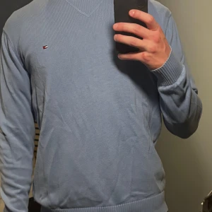 Ljusblå stickad tröja Tommy Hilfiger - Säljer en ljusblå stickad tröja från Tommy Hilfiger med klassisk v-ringning och diskret logga på bröstet. Tröjan har ribbade muddar och är långärmad, perfekt för lager-på-lager under kyliga dagar.