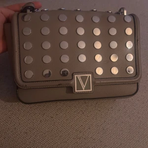 Axelväska från Victoria's Secret med nitar - Cool beige axelväska från Victoria's Secret i skinnimitation med stora silvriga nitar på locket och kedjerem i metall. Väskan har ett stilrent rutigt foder och en praktisk ficka på baksidan. Perfekt för dig som vill sticka ut med en edgy accessoar.