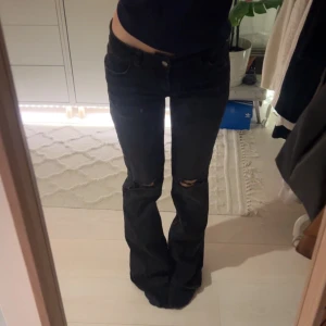 Svarta bootcut jeans från Nelly - Svarta lågmidjade jeans från Nelly med bootcut passform och hål vid knäna. Jeans har broderi på bakfickorna och har stretchigt material. Storlek 36, jag är 165 och de är lite långa på mig. Jag har använt dem kanske 2 gånger så väldigt bra skick.