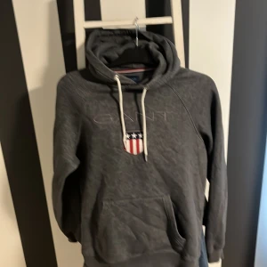 Mörkgrå hoodie från GANT med tryck - Mörkgrå hoodie från GANT med broderad logga och amerikanskt sköldtryck på bröstet. Tröjan har huva med vita snören, känguruficka och långa ärmar. Perfekt för dig som gillar klassisk streetstyle och vill ha något bekvämt och snyggt.