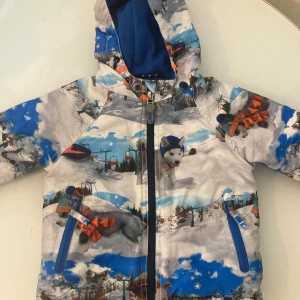 Färgglad vinterjacka från Next- FRI FRAKT - Säljer en cool vinterjacka från Next med unikt tryck av snölandskap, hundar med mössa och liftar. Jackan har huva med blå fleecefoder, två fickor med blå dragkedjor och hel dragkedja framtill. Jackan är fodrade för extra värme.