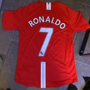 Manchester United Ronaldo 7 Nike tröja - Säljer en klassisk Manchester United fotbollströja med Ronaldo och nummer 7 på ryggen. Tröjan är röd med vita detaljer, har korta ärmar och är tillverkad i lätt, ventilerande polyester. Framsidan har AIG-logga och klubbmärke, perfekt för dig som älskar fotboll och United.