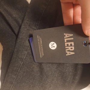 Svarta jeans från ALERA, storlek M - Svarta jeans från ALERA i klassisk rak modell. Jeansen har en snygg mörk tvätt och är tillverkade i denim. Bakfickor och tydlig ALERA-patch baktill i midjan. Perfekta för dig som gillar stilrena och enkla jeans.