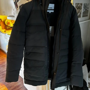 Helt ny jacka storlek S - Svart pufferjacka från Just Junkies med huva, stora fickor med lock och dragkedja framtill. Jackan är vadderad och har en clean, stilren look med raka linjer och quiltad design. Perfekt för kalla dagar när du vill hålla värmen och ändå se snygg ut.