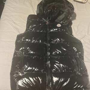 Svart dunväst från Moncler med huva - Snygg svart dunväst från Moncler med glansig finish och huva. Västen har två fickor med dragkedja och klassisk Moncler-logga på bröstet. Perfekt för lager-på-lager och håller dig varm under kyliga dagar.