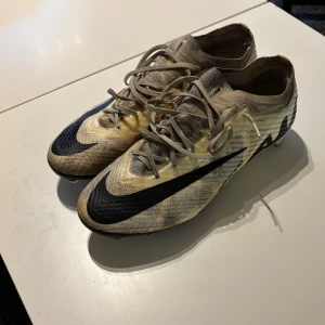 Nike Mercurial fotbollsskor beige/svart - Säljer ett par Nike Mercurial Zoom fotbollsskor i beige och svart med stora svarta swoosh-loggor. Skorna har snörning, låg profil och ovandel i syntetmaterial för lätt känsla och bra bollkontroll. Sulan har dobbar anpassade för spel på naturgräs.