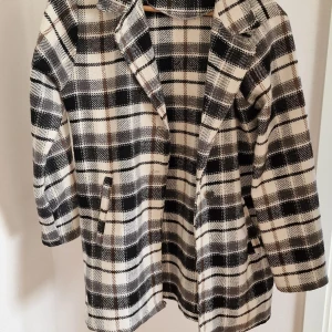 Rutig svartvit kappa med krage - Snygg rutig kappa i svart, grått och vitt med klassisk krage och raka ärmar. Jackan har två fickor framtill och är i ett mjukt, lite tjockare material som passar perfekt till höst och vinter. Stängs framtill och har en stilren, tidlös look.
