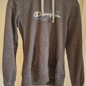 Marinblå hoodie från Champion med tryck - Mysig marinblå hoodie från Champion med vit dragsko och klassiskt tryck på bröstet. Tröjan har känguruficka framtill och ribbade muddar vid ärmslut och nederkant. Perfekt för chill dagar eller när du vill ha en sportig look.