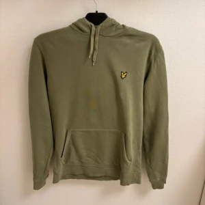 Olivgrön hoodie från Lyle & Scott - Olivgrön hoodie från Lyle & Scott med klassisk gul logga på bröstet. Tröjan har huva med snörning, magficka och långa ärmar. Mjuk bomullskvalitet och relaxed passform, perfekt för chill dagar. Snygg och enkel att matcha med jeans eller joggers.