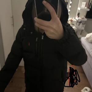 Svart dunjacka från Ralph Lauren - Säljer en svart dunjacka från Ralph Lauren med huva och dragkedja framtill. Jackan har en röd broderad logga på bröstet och praktiska fickor med dragkedja. Perfekt för kalla dagar och riktigt snygg streetstil.