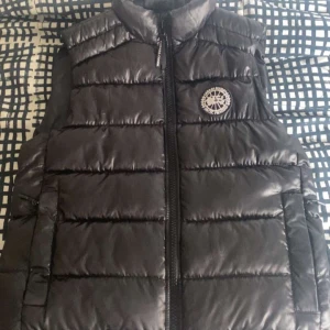 Svart dunväst från Canada Goose - Snygg svart dunväst från Canada Goose med quiltad design och hög krage. Västen har dragkedja framtill, två sidofickor med dragkedja och ett broderat logomärke på bröstet. Perfekt för lager-på-lager under kyliga dagar.