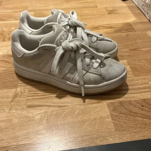 Adidas Campus sneakers i ljusgrå mocka - Säljer ett par Adidas Campus sneakers i ljusgrå mocka med klassiska vita ränder på sidorna och vit sula. Skorna har rund tå, snörning och Adidas logga på plösen. Perfekta för dig som gillar en clean och stilren look. Säljer mina favoriter då dom är för små .jag köpte för 1300