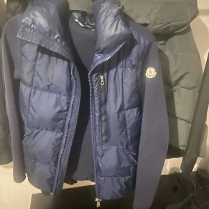 Mörkblå dunjacka från Moncler - Snygg mörkblå dunjacka från Moncler med stickade ärmar och vadderad framsida. Jackan har dragkedja, huva och klassisk Moncler-logga på ärmen. Perfekt för kyliga dagar och ger en sportig men clean look.
