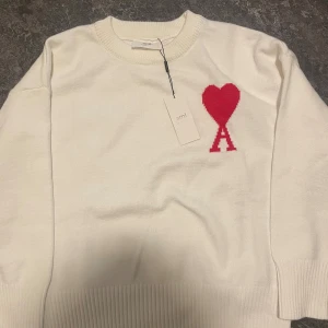 Vit stickad tröja från Ami - Vit stickad tröja från Ami med rund hals och ribbade muddar. På bröstet finns ett rött hjärta med bokstaven A, som är typiskt för märket. Tröjan har lång ärm och är gjord i mjukt material som känns skönt mot huden. Perfekt för dig som gillar stilrena detaljer.