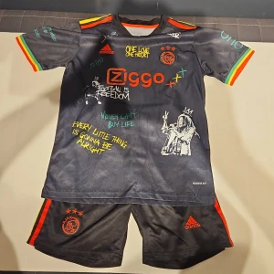 Ajax Bob Marley fotbollströja Adidas - Ajax x Bob Marley limited edition fotbollströja från Adidas. Svart med färgglada detaljer i rött, gult och grönt på ärmarna, tryckta citat och illustrationer inspirerade av Bob Marley. Kortärmad, med Ajax-emblem och Ziggo sponsortryck. Material: polyester, Aeroready. Jag köpte den på en marknad i Spanien så det är en replika.