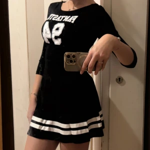 Svart sportig kortklänning XSS-XS - Sportig svart klänning med vita tryckdetaljer och texten 'FANTASTIC 94' framtill. Klänningen har trekvartsärm, rund halsringning och två vita ränder längst ner på kjolen. Sitter snyggt och har en ungdomlig vibe.