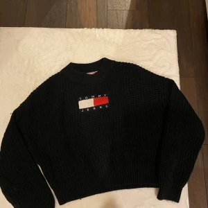 Svart stickad tröja Tommy Jeans - Svart stickad tröja från Tommy  Hilfiger med broderad logga i vitt och rött framtill. Tröjan har rund hals, ribbade muddar och en loose passform. Perfekt för dig som gillar klassisk streetstyle med en modern touch.