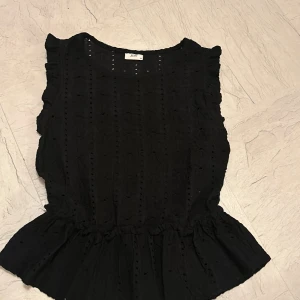 Svart blus med volang från JDY - Supersöt svart blus från JDY i storlek XS. Blusen har broderade detaljer, volang vid axlarna och en lätt rynkad midja som ger en feminin siluett. Ärmlös modell med rund halsringning och snygg struktur i tyget.