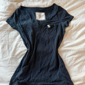 Mörkblå t-shirt Abercrombie & Fitch XS - Mörkblå t-shirt från Abercrombie & Fitch med v-ringning och liten bröstficka med broderad logga. Klassisk passform och korta ärmar, perfekt till jeans eller shorts. i storlek xs så den har en tajt passform,använd ett par gånger men köpt på second hand