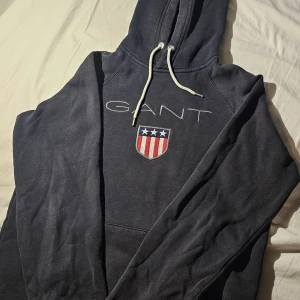 Mörkblå hoodie från GANT med logga - Säljer en mörkblå hoodie från GANT med broderad logga och sköld på bröstet. Tröjan har huva med vita snören och en stor magficka. Perfekt för dig som gillar klassisk och sportig stil.