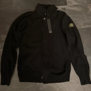 Svart stickad kofta från Stone Island - Svart stickad kofta från Stone Island med klassisk logotyp på ärmen. Koftan har hög krage med knappdetaljer, ribbade muddar och knäppning framtill. Perfekt för dig som gillar stilrena och exklusiva plagg med streetkänsla. Satt stor för mig, anledning till att den är till salu!