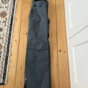 Grå täckbyxor från Stellar Equipment - Snygga grå skalbyxor från Stellar Equipment med flera dragkedjeförsedda fickor, förstärkta benslut och justerbar midja. Byxorna har en clean look och är perfekta för vinterns äventyr. Materialet är slitstarkt och vattenavvisande syntet.