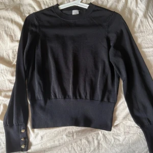 Svart stickad tröja från H&M - Svart stickad tröja från H&M med långärmade puffärmar och bred ribbad kant nedtill. Snygga guldfärgade knappar på ärmsluten ger en extra detalj. Perfekt för dig som gillar stilrena plagg med lite edge.