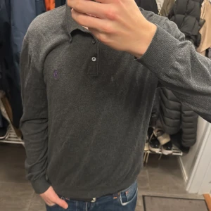 Mörkgrå pikétröja från Polo Ralph Lauren - Snygg mörkgrå långärmad pikétröja från Polo Ralph Lauren i mjuk pima bomull. Klassisk krage, två knappar och diskret lila logga broderad på bröstet. Perfekt för dig som gillar stilrena och bekväma plagg med premiumkänsla.