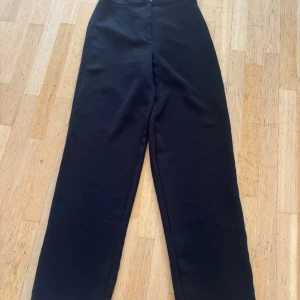 Svarta kostymbyxor med hög midja - Stilrena svarta kostymbyxor med hög midja och raka ben. Byxorna har en enkel design utan synliga fickor eller detaljer, vilket ger en clean och modern look. Perfekta för dig som gillar minimalistisk stil och vill ha ett par klassiska svarta byxor.