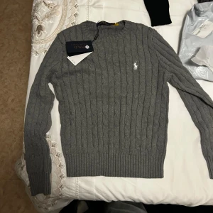 Kabelstickad tröja Polo Ralph Lauren - Kabelstickad tröja från Polo Ralph Lauren i grått och ljuslila. Klassisk rund halsringning, ribbade muddar och broderad logga på bröstet. Perfekt för lager-på-lager och snygg till jeans eller kjol. Priset kan diskuteras 