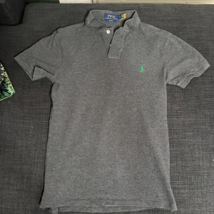 Grå pikétröja Polo Ralph Lauren - Snygg grå pikétröja från Polo Ralph Lauren i slim fit-modell. Klassisk krage, korta ärmar och två knappar framtill. Ikonisk grön logga broderad på bröstet. Perfekt för dig som gillar stilrena och tidlösa plagg. Knappt använd.