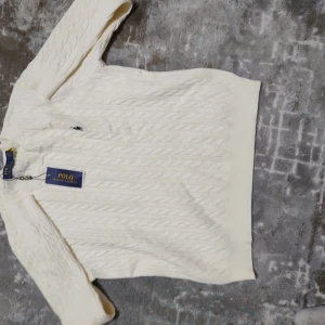 Vit kabelstickad tröja Polo Ralph Lauren - Kabelstickad vit tröja från Polo Ralph Lauren med klassisk rund halsringning och broderad logga på bröstet. Tröjan har långa ärmar och ribbade muddar för en snygg och tidlös look. Perfekt för lager-på-lager under höst och vinter.