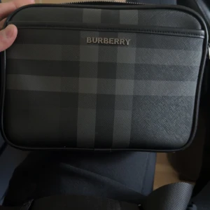 Svart rutig axelväska från Burberry - Snygg svart axelväska från Burberry med klassiskt rutigt mönster i mörka toner. Väskan har justerbar axelrem och dragkedja upptill. Tillverkad i skinn med stilren metallogo framtill och praktiskt innerfack. Perfekt för dig som vill ha en clean och lyxig look.